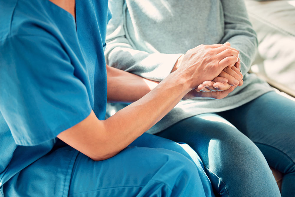Healthcare professional in blue scrubs compassionately holding hands with a seated patient in gray clothing.